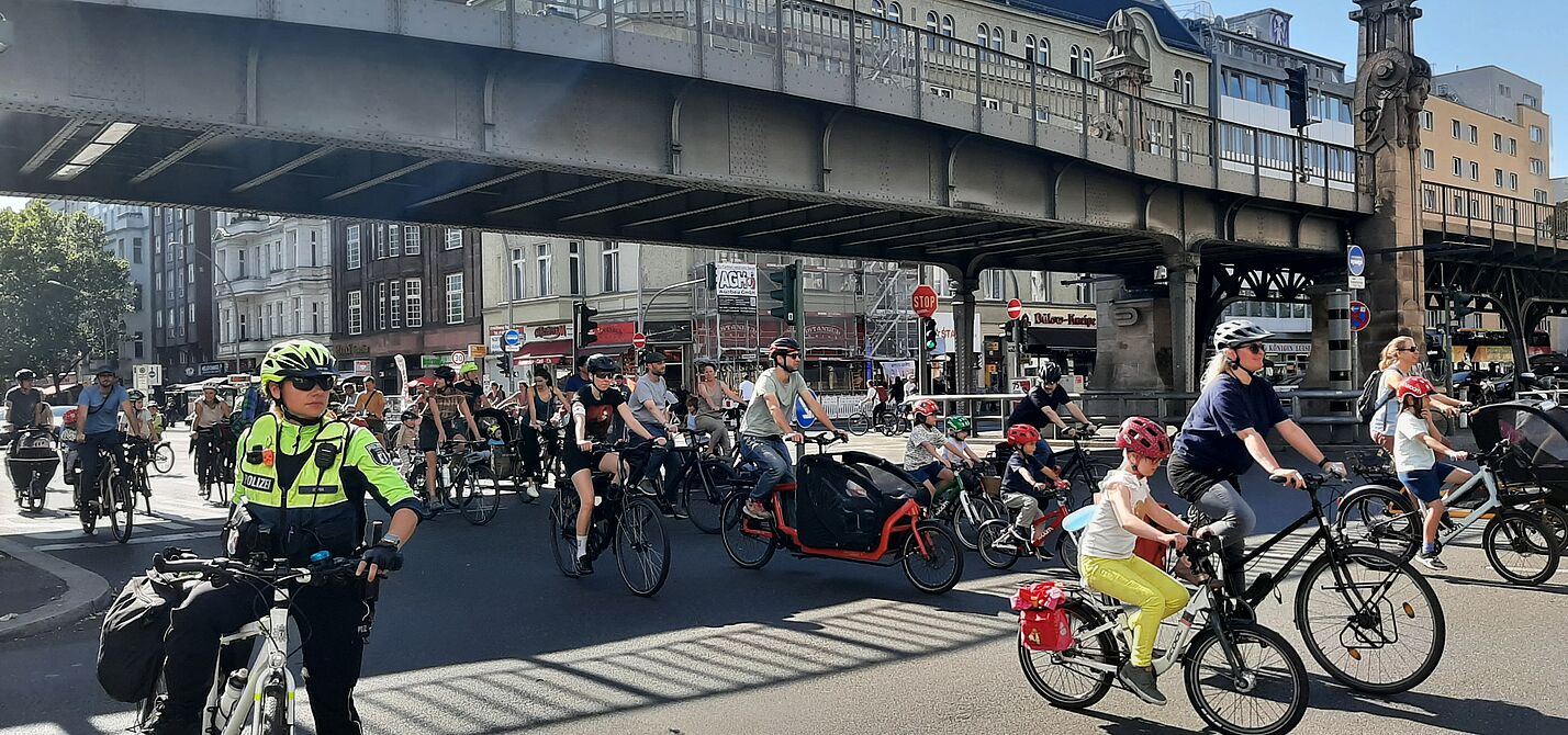 Eine Gruppe Erwachsener und Kinder fahren auf ihren Fahrrädern unter einer Brücke auf der Fahrbahn der Bülowstraße in Schönberg. Im Vordergrund ist eine Fahrradpolizistin zu sehen.