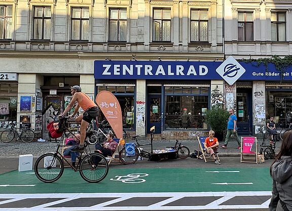 Ein Zweirad fährt über einen grünen Pop-Up Radweg, Menschen auf Liegestühlen im Hintergrund