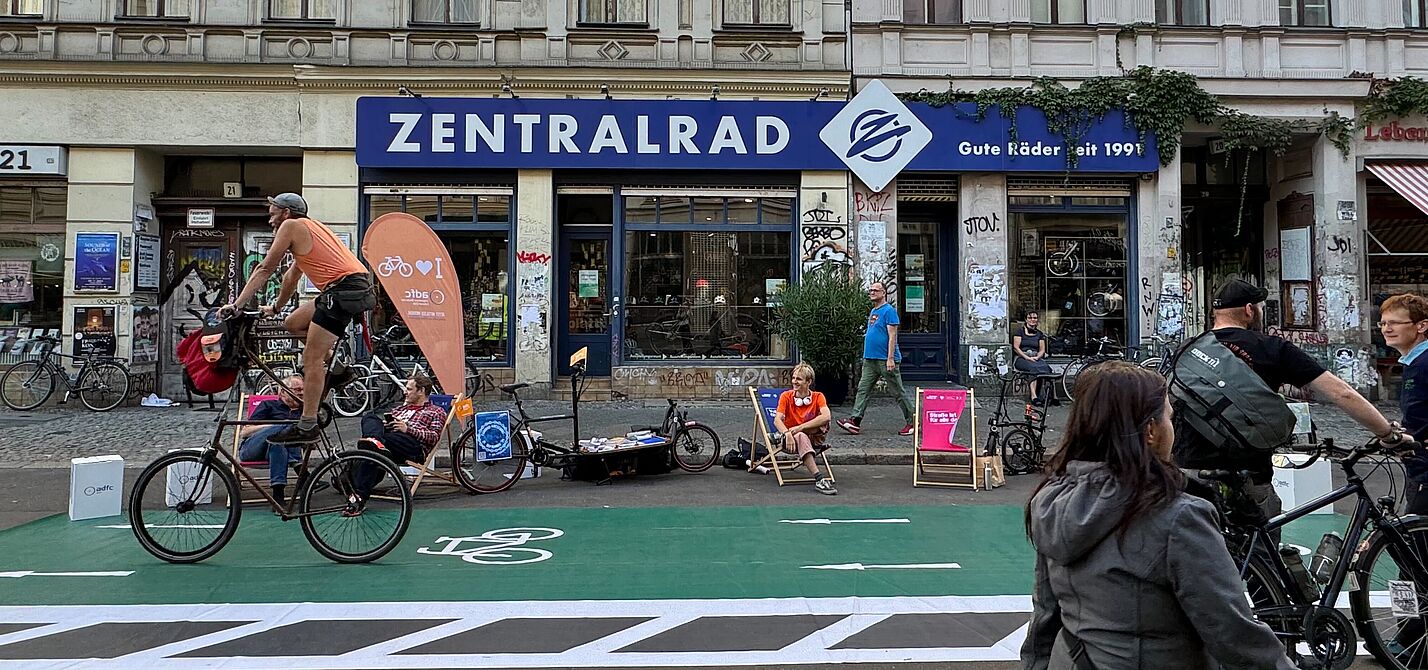 PARKing Day 2024 ADFC Berlin Ein Zweirad fährt über einen grünen Pop-Up Radweg, Menschen auf Liegestühlen im Hintergrund