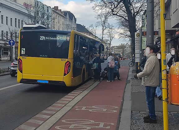 Das Bild zeigt im rechten Bereich eine Haltestelle, daneben ein Haltestellenkap mit darüber geführten Radweg und ein haltender Bus. Fahrgäste steigen ein und aus. 