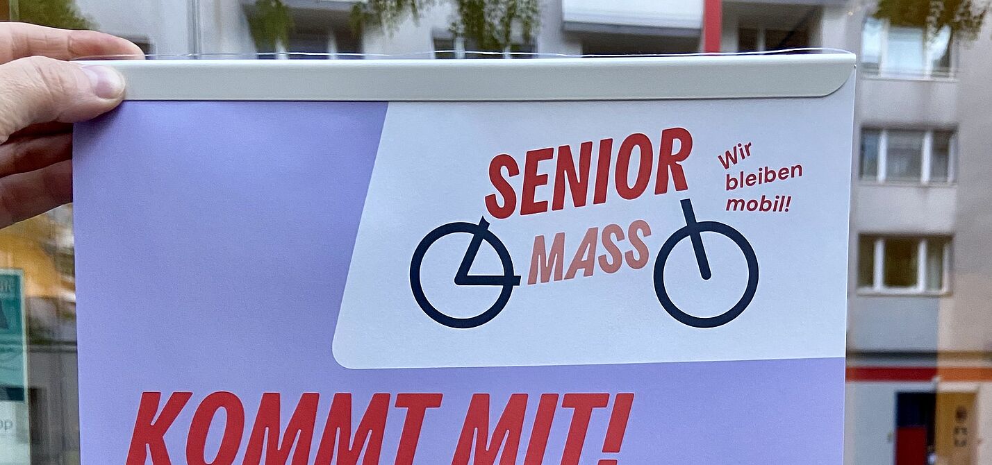 Senior Mass Plakat von Changing Cities Ein Plakat mit dem Titel „Senior Mass – Kommt mit!“ wird vor einer Schaufensterscheibe ins Licht gehalten.