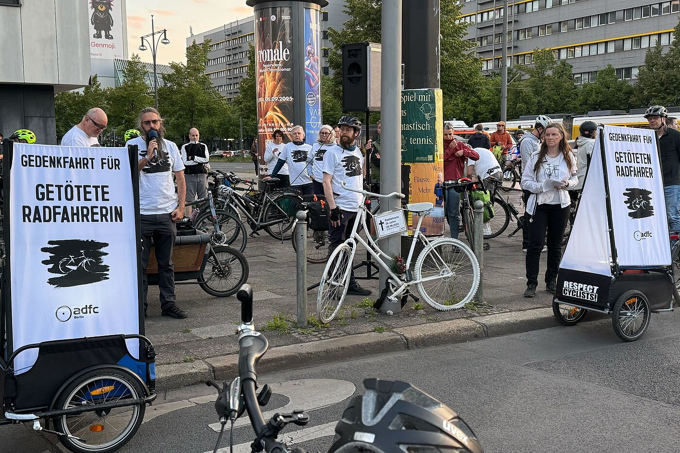 Menschen halten am Straßenrand eine Rede zwischen zwei Schildern "Gedenkfahrt für getötete Radfahrerin"