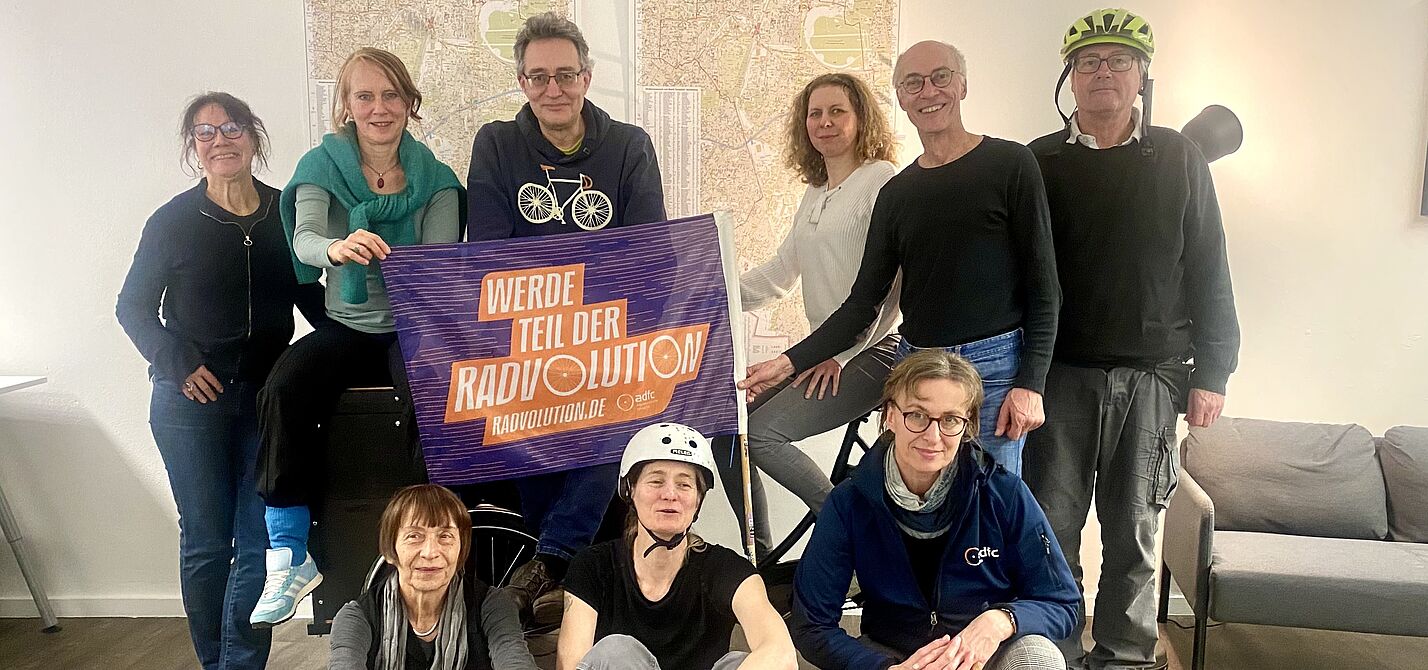 Stadtteilgruppe Schöneberg im Raum der Begegnung Dominicusstraße Eine Gruppe Menschen stehen um und sitzen vor und auf einem Lastenrad, einige tragen Fahrradhelme. Eine ADFC Fahne mit Schrift "Werde Teil der Radvolution" ist im Mittelpunkt. Im Hintergrund sind Karten von Berlin Schöneberg sichtbar, aber nicht zu entziffern.
