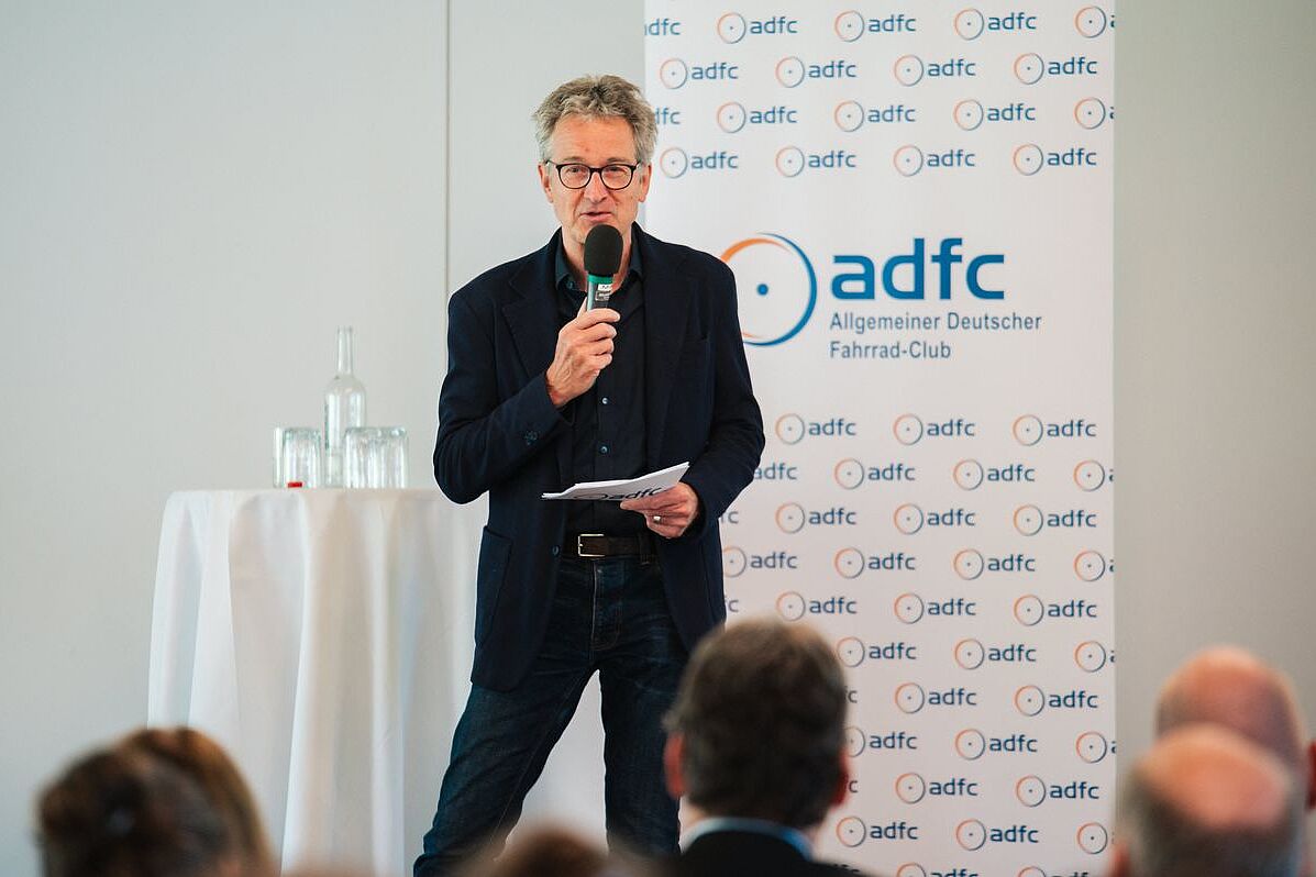 ADFC-Bundesvorstandsmitglied Christian Tänzler steht mit Mikrofon auf der Bühne des ADFC-Radtourismuskongresses. Im Hintergund eine weiße Wand und ein weißer Aufsteller mit ADFC-Logos.