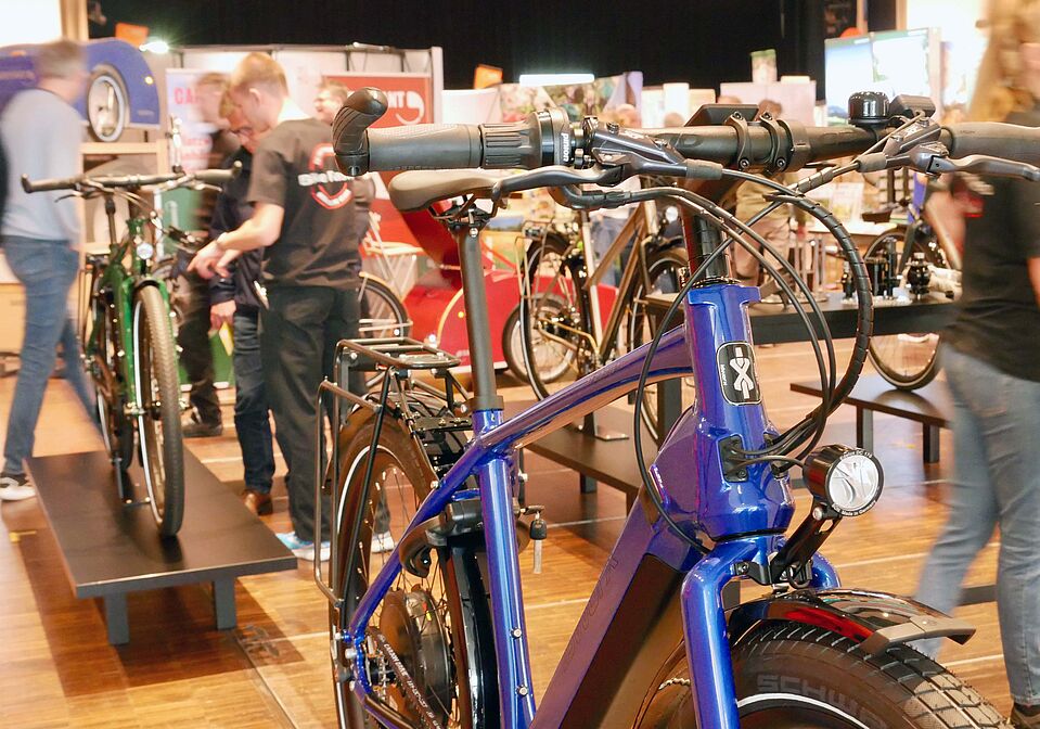 Messe rad+freizeit 2025 Nahaufnahme eines blauen Elektrofahrrads auf einer Messe. Der Fokus liegt auf dem Lenker, dem Frontscheinwerfer und dem Rahmen. Im Hintergrund sind unscharf weitere Fahrräder und Besuchende zu erkennen, die sich an Ständen informieren.