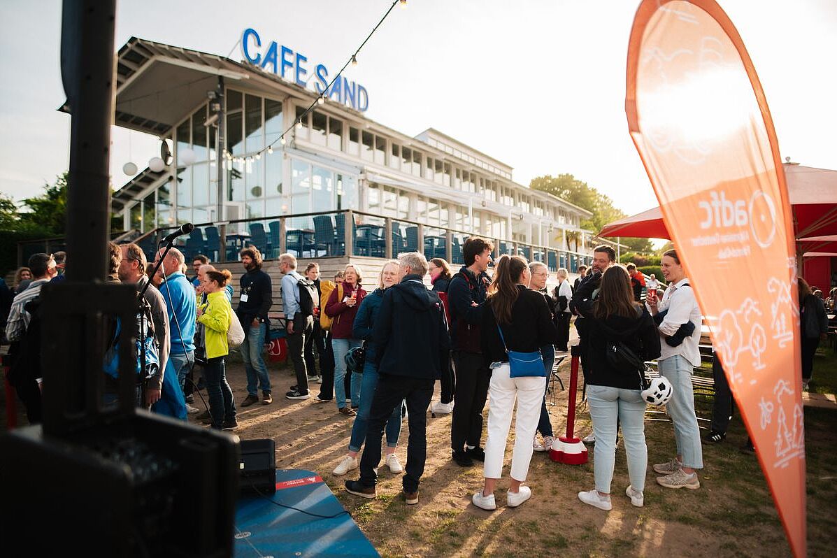 Abendstimmung am Weserstrand. zahlreiche menschen vor dem Café Sand in Bremen in Gesprächen. Im Vordergrund eine orangefarbene ADFC-Beachflag. 